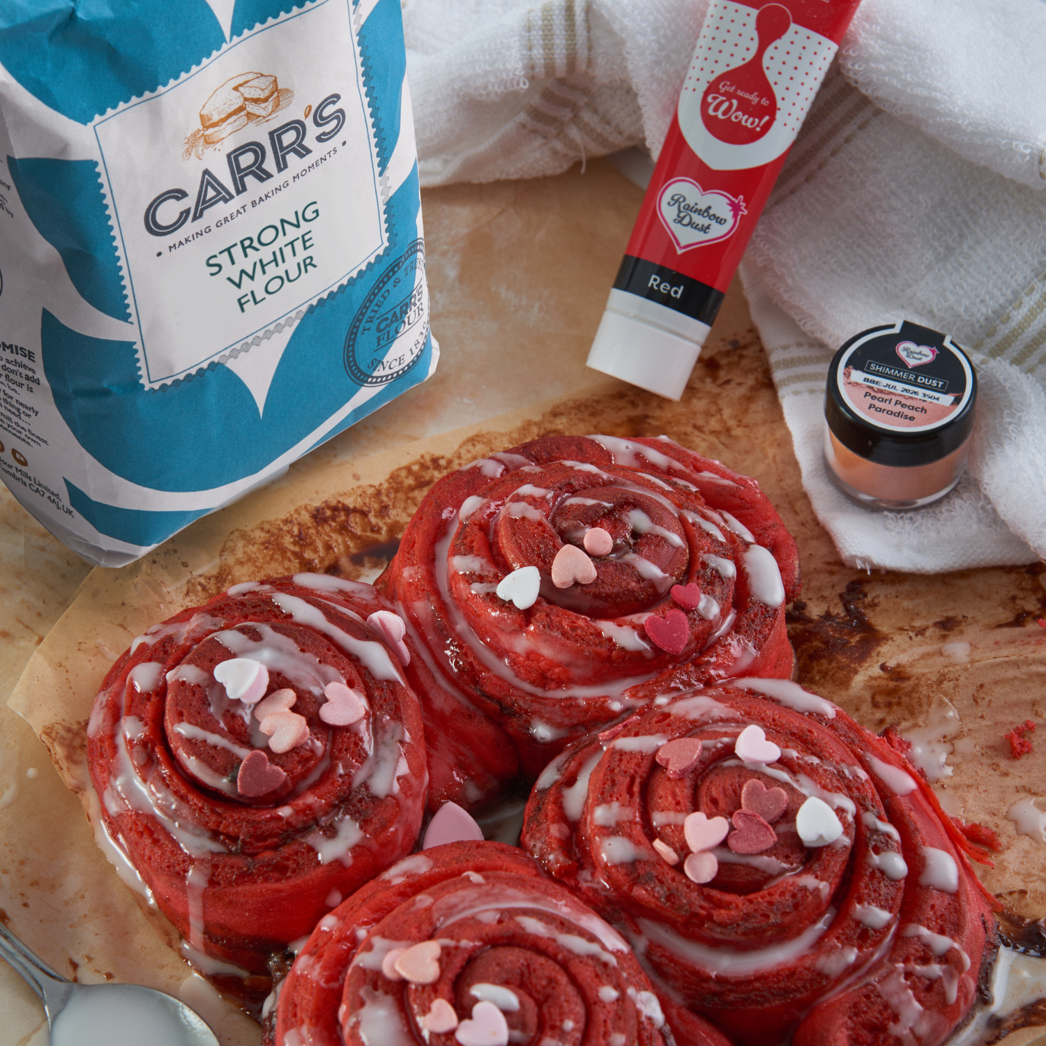 Red Valentine's cinnamon buns topped with heart sprinkles beside baking ingredients