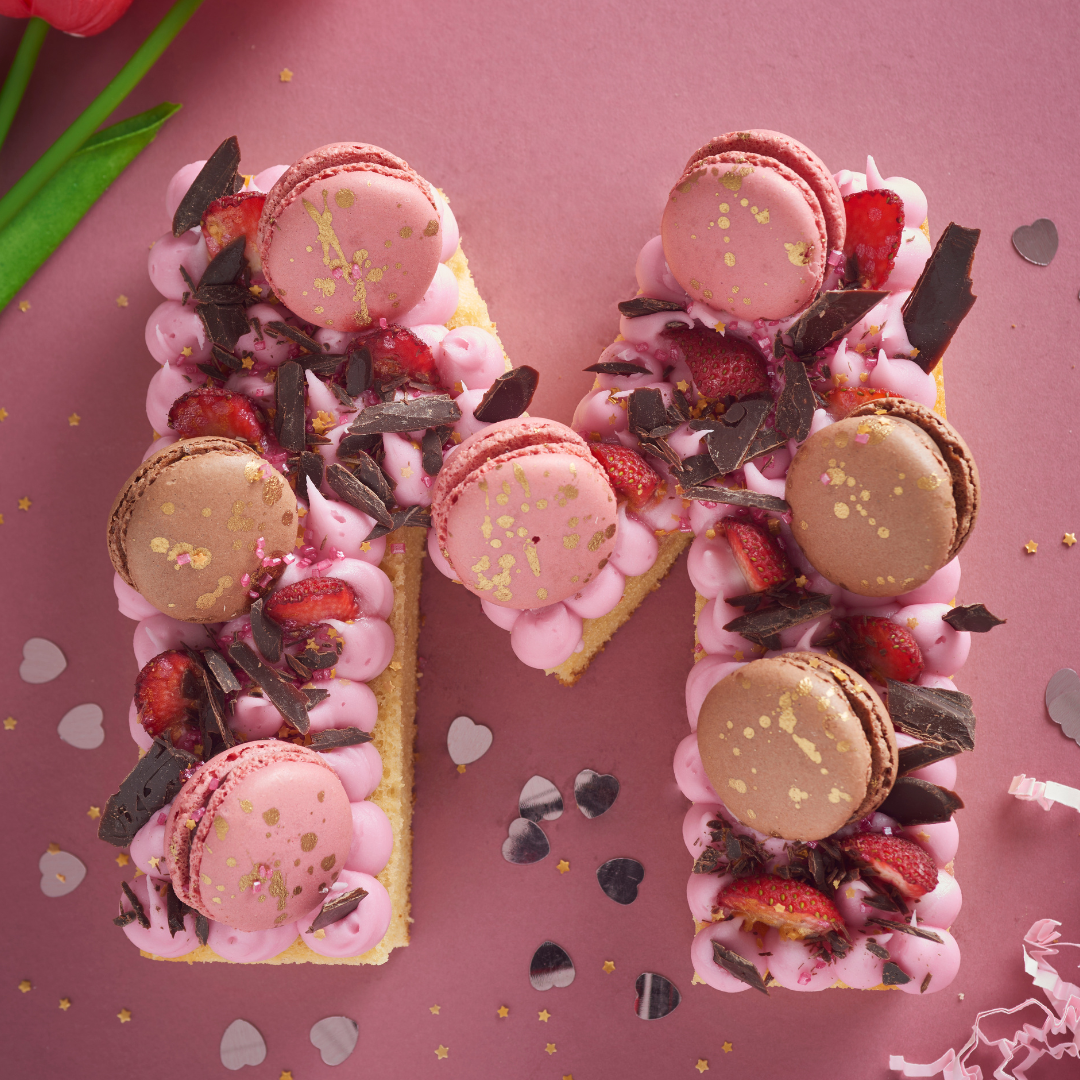 Pink letter M cake topped with macarons, berries and gold sprinkles